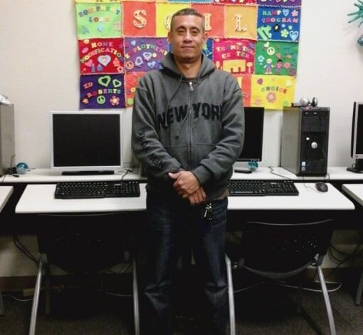 Young man standing in front of computer classroom wearing a dark gray hoodie sweatshirt with a patchwork colorful quilt hanging on the wall behind him.