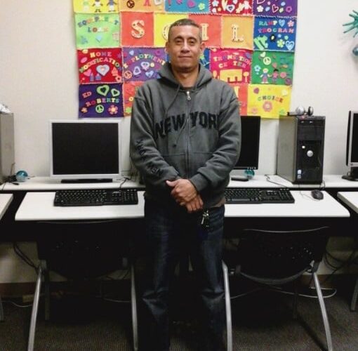 Young man standing in front of computer classroom wearing a dark gray hoodie sweatshirt with a patchwork colorful quilt hanging on the wall behind him.