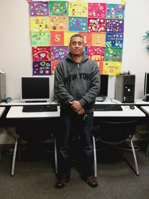 Young man standing in front of computer classroom wearing a dark gray hoodie sweatshirt with a patchwork colorful quilt hanging on the wall behind him.