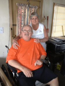 Man in orange shirt sitting in a wheelchair with a woman in a white blouse standing behind him with her hands on his shoulders.