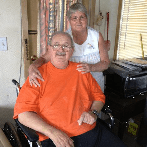 Man in orange shirt sitting in a wheelchair with a woman in a white blouse standing behind him with her hands on his shoulders.