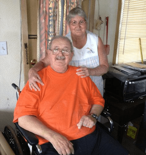 Man in orange shirt sitting in a wheelchair with a woman in a white blouse standing behind him with her hands on his shoulders.